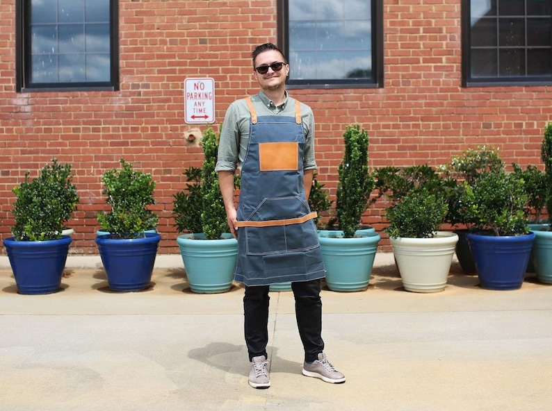 May include: A man wearing a blue denim apron with brown leather straps and a brown leather patch pocket. He is standing in front of a brick wall with potted plants.