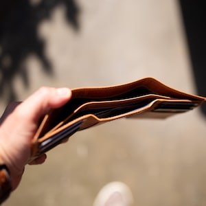 May include: Open, light brown leather wallet with multiple card slots and bill compartments. The wallet is held open, revealing the interior. A brown leather watch is visible on the wrist.