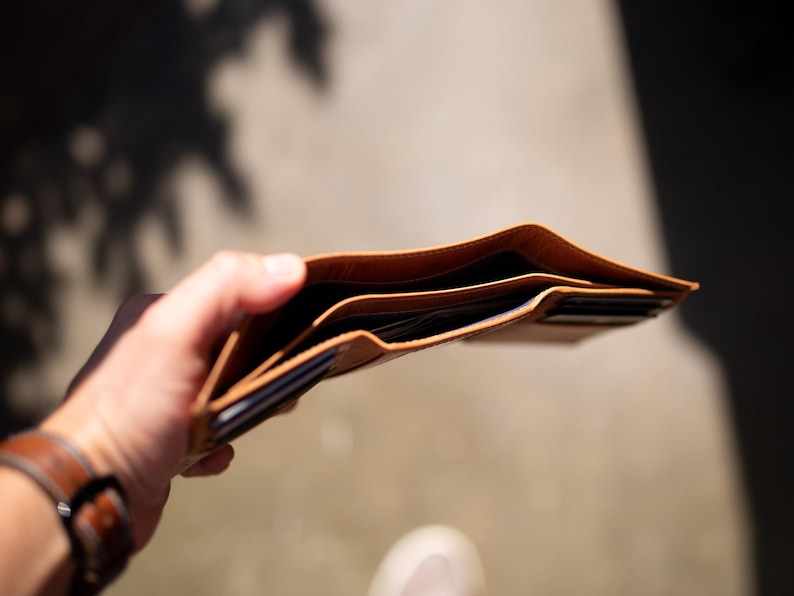 May include: Open, light brown leather wallet with multiple card slots and bill compartments. The wallet is held open, revealing the interior. A brown leather watch is visible on the wrist.