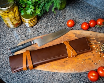 May include: A brown leather knife roll with tan leather straps and buckles, wrapped around a long, black-handled chef's knife on a wooden cutting board.