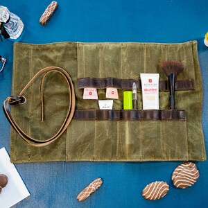 K&ouml;nnte beinhalten: Eine gr&uuml;ne Canvas-Rolltasche mit braunen Lederriemen und Taschen. Die Tasche ist ge&ouml;ffnet und enth&auml;lt verschiedene Make-up- und Beauty-Produkte, darunter einen Pinsel, eine Tube BB-Creme und einen Lipgloss.