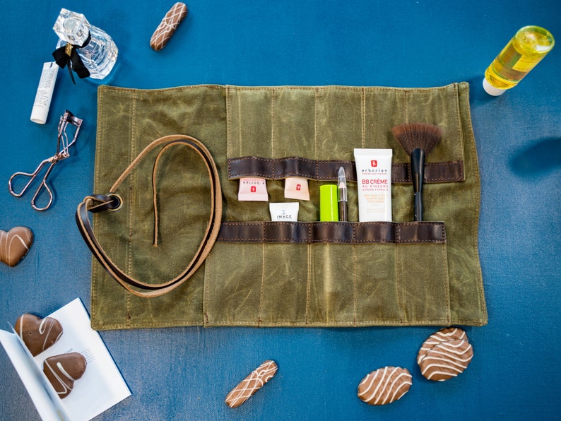 K&ouml;nnte beinhalten: Eine gr&uuml;ne Canvas-Rolltasche mit braunen Lederriemen und Taschen. Die Tasche ist ge&ouml;ffnet und enth&auml;lt verschiedene Make-up- und Beauty-Produkte, darunter einen Pinsel, eine Tube BB-Creme und einen Lipgloss.