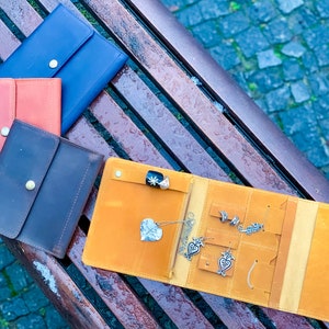 May include: An open, mustard-yellow leather jewelry organizer with various silver earrings and a ring. Several closed leather wallets in blue, orange, and brown are also visible. The items are arranged on a wooden bench.