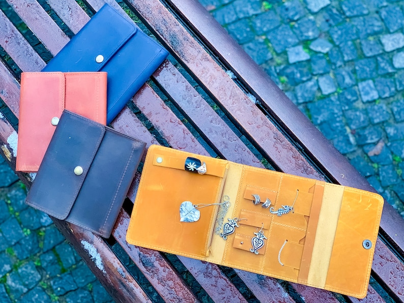 May include: An open, mustard-yellow leather jewelry organizer with various silver earrings and a ring. Several closed leather wallets in blue, orange, and brown are also visible. The items are arranged on a wooden bench.