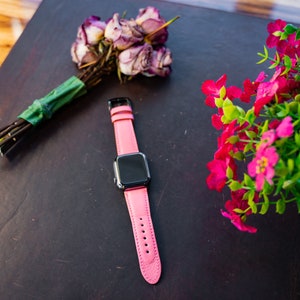 Puede incluir: Una correa de reloj de cuero rosa con una esfera negra. La correa est&aacute; sobre una superficie marr&oacute;n oscura con un ramo de rosas rosas y una planta en maceta con flores rosas.
