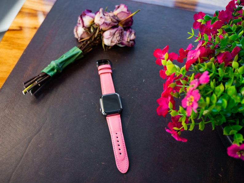 Puede incluir: Una correa de reloj de cuero rosa con una esfera negra. La correa est&aacute; sobre una superficie marr&oacute;n oscura con un ramo de rosas rosas y una planta en maceta con flores rosas.