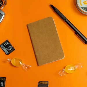 May include: A brown leather notebook with a black pen, two memory cards, two yellow candies, and a small container of leather cream on an orange surface.
