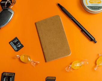 May include: A brown leather notebook with a black pen, two memory cards, two yellow candies, and a small container of leather cream on an orange surface.