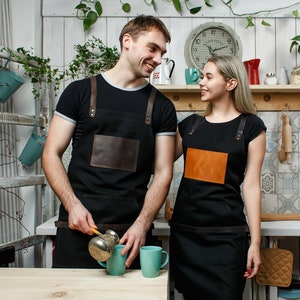 May include: Two people wearing black aprons with leather accents stand in a kitchen. The person on the left is pouring coffee from a cezve into a blue mug. The person on the right is smiling and looking at the person on the left. There are two blue mugs on a wooden table.