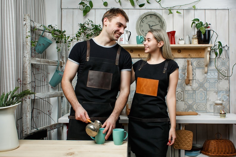 May include: Two people wearing black aprons with leather accents stand in a kitchen. The person on the left is pouring coffee from a cezve into a blue mug. The person on the right is smiling and looking at the person on the left. There are two blue mugs on a wooden table.