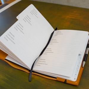 May include: An open brown leather bound notebook with a black ribbon bookmark. The pages are lined and have headings for "Website", "Username", "Password/hint", and "Notes". A silver pen is tucked into the binding.