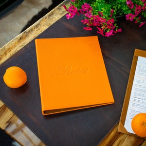 May include: A brown wooden table with a dark brown leather placemat. On the placemat is a bright orange menu with the word "Bistro" embossed in white. There is a small orange on the placemat and a menu with the word "Lunch" on the table.