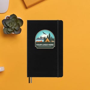 Puede incluir: Cuaderno negro con cierre elástico negro y una pegatina colorida con una escena de camping y el texto "YOUR LOGO HERE".