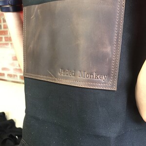 May include: A black canvas apron with a brown leather patch that says "Jaded Monkey" in white stitching.