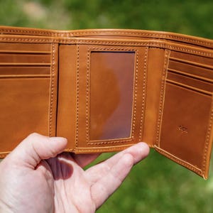 May include: Open, tri-fold brown leather wallet with multiple card slots and a clear ID window. The wallet is held open, showcasing the interior design and stitching details. The background is a blurred green.