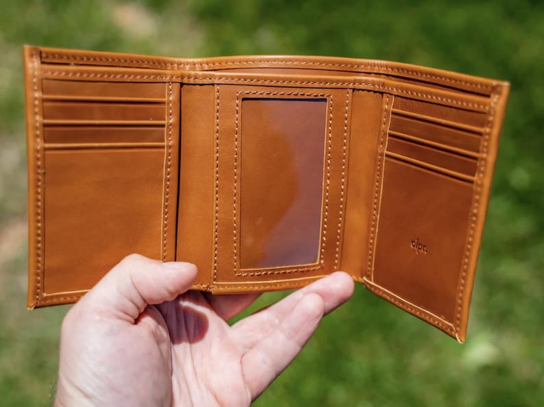 May include: Open, tri-fold brown leather wallet with multiple card slots and a clear ID window. The wallet is held open, showcasing the interior design and stitching details. The background is a blurred green.
