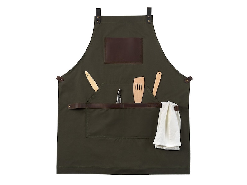 May include: A green canvas apron with brown leather straps and a large pocket. The pocket has a brown leather flap and is filled with a wooden spatula, a wooden spoon, and a small knife.