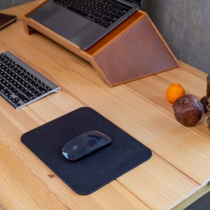 May include: A wooden desk with a black mouse pad and a wireless mouse. A keyboard, a laptop, a notebook, a pen, a pine cone, a pomegranate, and an orange are also on the desk.