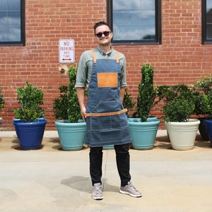 May include: A man wearing a blue denim apron with brown leather straps and a brown leather patch pocket. He is standing in front of a brick wall with potted plants.