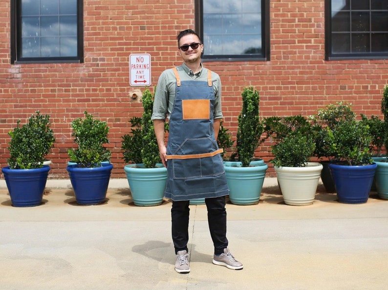 May include: A man wearing a blue denim apron with brown leather straps and a brown leather patch pocket. He is standing in front of a brick wall with potted plants.
