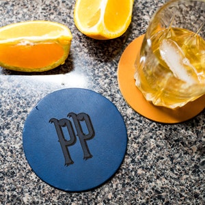 Könnte beinhalten: Zwei Lederuntersetzer, einer blau mit den Initialen "PP" in Schwarz und einer orange. Ein Glas Whisky mit Eis steht auf dem orangen Untersetzer.
