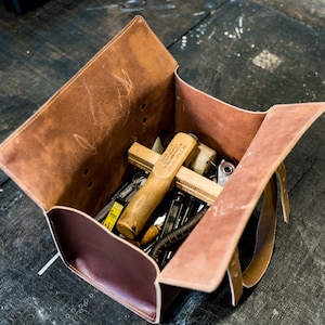K&ouml;nnte beinhalten: Eine braune Ledertasche f&uuml;r Werkzeuge mit offenem Klappdeckel, die eine Vielzahl von Werkzeugen enth&uuml;llt, darunter einen Holzhammer, ein Lineal und einen Schraubenschl&uuml;ssel aus Metall.