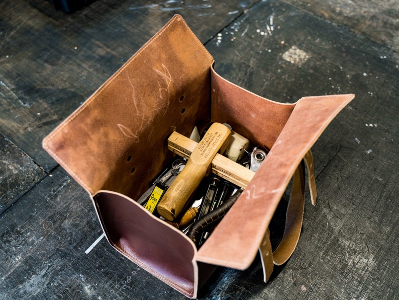 K&ouml;nnte beinhalten: Eine braune Ledertasche f&uuml;r Werkzeuge mit offenem Klappdeckel, die eine Vielzahl von Werkzeugen enth&uuml;llt, darunter einen Holzhammer, ein Lineal und einen Schraubenschl&uuml;ssel aus Metall.