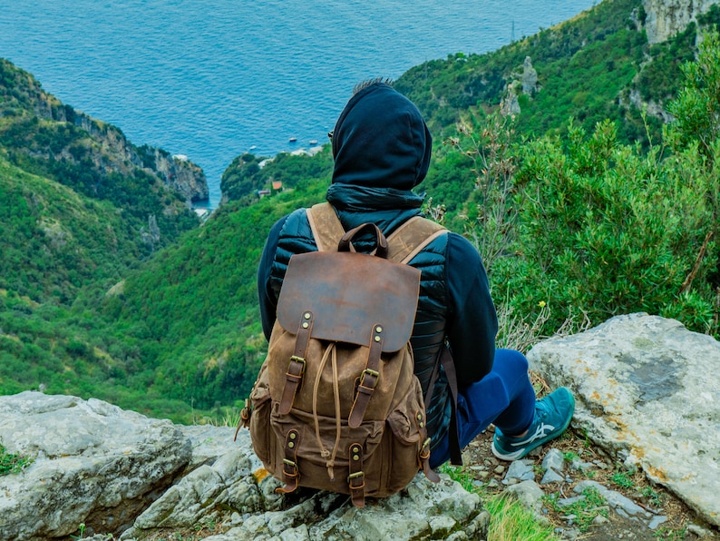 Puede incluir: Una mochila de lona y cuero marr&oacute;n con m&uacute;ltiples bolsillos y correas. La mochila tiene un cierre de solapa y una parte superior con cord&oacute;n. La persona lleva una chaqueta negra y pantalones azules, sentada en una roca con vistas a un valle verde y el mar.