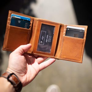 May include: Open, tri-fold leather wallet in a warm brown hue, showcasing multiple card slots. The wallet is held open, revealing credit cards and a business card. The stitching is visible, and the wallet appears to be well-crafted.