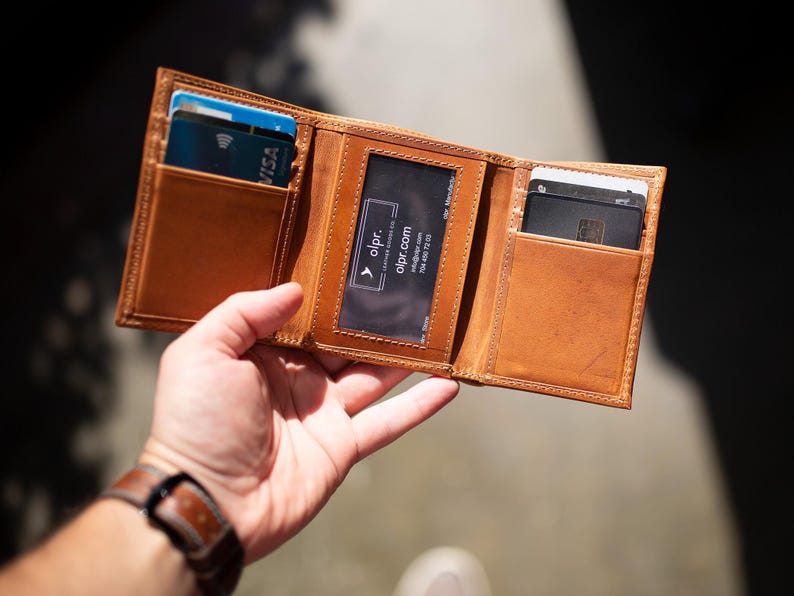 May include: Open, tri-fold leather wallet in a warm brown hue, showcasing multiple card slots. The wallet is held open, revealing credit cards and a business card. The stitching is visible, and the wallet appears to be well-crafted.
