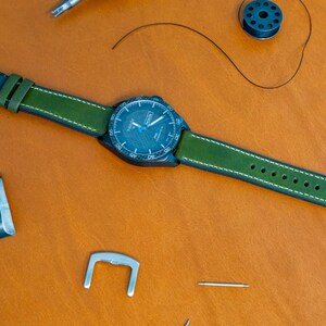 Puede incluir: Un reloj de pulsera con esfera azul oscuro y correa de cuero verde con costuras blancas. El reloj est&aacute; sobre una superficie de cuero marr&oacute;n, con herramientas de relojero cerca.