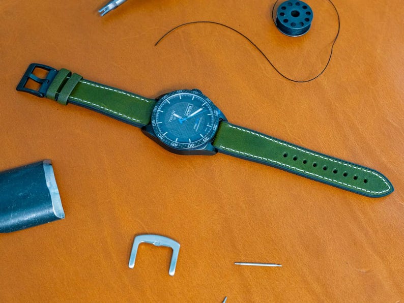 Puede incluir: Un reloj de pulsera con esfera azul oscuro y correa de cuero verde con costuras blancas. El reloj est&aacute; sobre una superficie de cuero marr&oacute;n, con herramientas de relojero cerca.