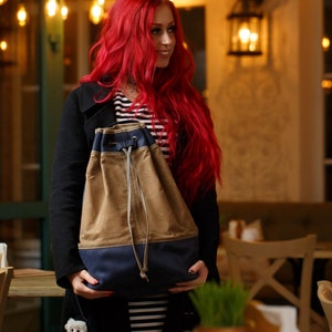 May include: A woman with long red hair is holding a brown and blue drawstring bag. The bag has a canvas-like texture and a contrasting blue bottom. She is standing in a restaurant setting.
