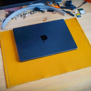 Könnte beinhalten: Eine rechteckige Schreibtischunterlage in Senfgelb mit strukturierter Oberfläche und genähten Kanten, auf der ein geschlossener dunkelgrauer Laptop mit einem schwarzen Apple-Logo in der Mitte des Deckels auf einer hellen Holzoberfläche liegt.