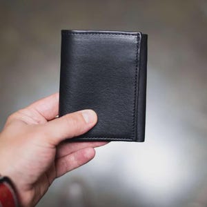 May include: A black leather wallet with visible stitching. The wallet is held in a hand, showcasing its compact, tri-fold design. The leather appears smooth and the wallet is closed.
