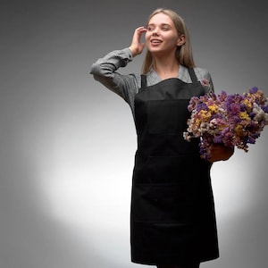 May include: A woman wearing a black apron and a plaid shirt holds a bouquet of dried flowers. The flowers are a mix of purple, pink, and white.