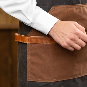 May include: A person wearing a brown and black apron with a leather belt and a brown pocket. They are holding a silver and black electric razor with a brush attachment.
