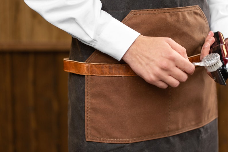 May include: A person wearing a brown and black apron with a leather belt and a brown pocket. They are holding a silver and black electric razor with a brush attachment.