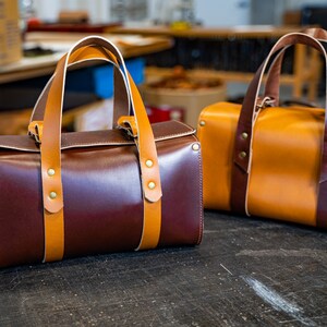 K&ouml;nnte beinhalten: Zwei Leder-Reisetaschen, eine braun und eine beige, mit braunen Lederriemen und Messing-Akzenten.