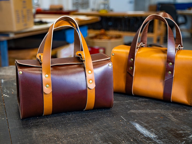 K&ouml;nnte beinhalten: Zwei Leder-Reisetaschen, eine braun und eine beige, mit braunen Lederriemen und Messing-Akzenten.