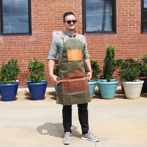May include: A man wearing a green canvas apron with brown leather accents and a brown leather patch on the front. He is standing in front of a brick wall with potted plants.