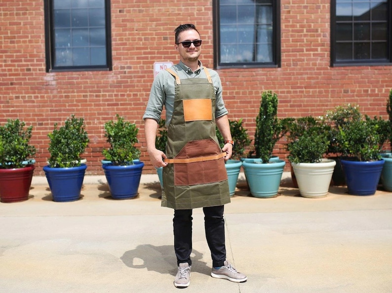 May include: A man wearing a green canvas apron with brown leather accents and a brown leather patch on the front. He is standing in front of a brick wall with potted plants.