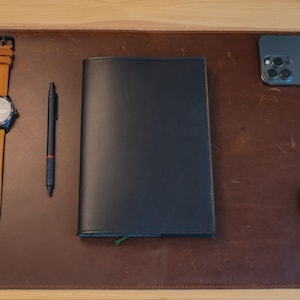 May include: A leather desk setup featuring a black notebook, a pen, a wristwatch with a tan leather band, a smartphone, and a computer mouse. The items are arranged on a brown leather desk mat.