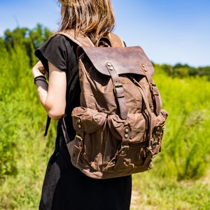Puede incluir: Mochila de lona marr&oacute;n con detalles de cuero. La mochila tiene m&uacute;ltiples bolsillos, correas ajustables y un cierre de solapa. La mochila es llevada por una persona con un vestido negro, sobre un fondo de follaje verde y cielo azul.