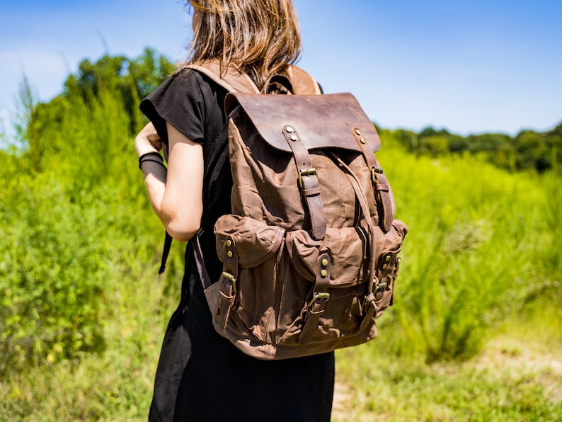 Puede incluir: Mochila de lona marr&oacute;n con detalles de cuero. La mochila tiene m&uacute;ltiples bolsillos, correas ajustables y un cierre de solapa. La mochila es llevada por una persona con un vestido negro, sobre un fondo de follaje verde y cielo azul.