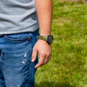 Puede incluir: Primer plano del brazo de una persona que lleva un reloj negro con una correa de cuero verde. El reloj tiene una esfera redonda. La persona lleva una camiseta gris y vaqueros azules.