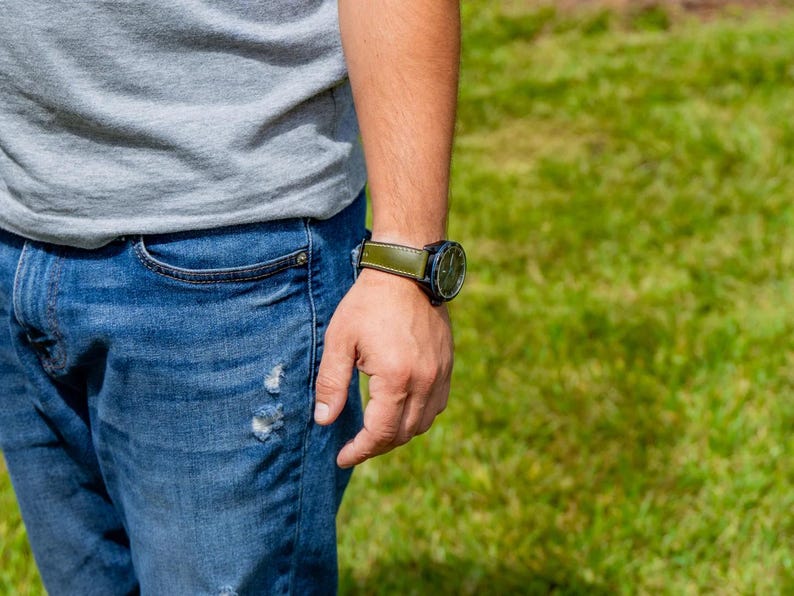 Puede incluir: Primer plano del brazo de una persona que lleva un reloj negro con una correa de cuero verde. El reloj tiene una esfera redonda. La persona lleva una camiseta gris y vaqueros azules.