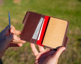 Könnte beinhalten: Ein braunes Leder-Notizbuchcover mit rotem Innenfutter und einer kleinen Tasche für Karten. Das Cover ist geöffnet und zeigt ein leeres Notizbuch mit linierten Seiten.