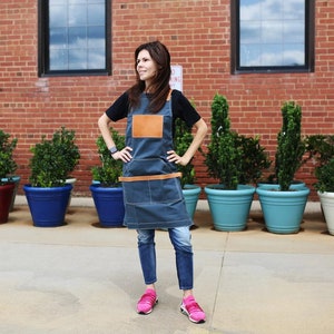 May include: A woman wearing a blue denim apron with a brown leather patch and a brown leather belt. She is standing in front of a brick wall with plants in pots.
