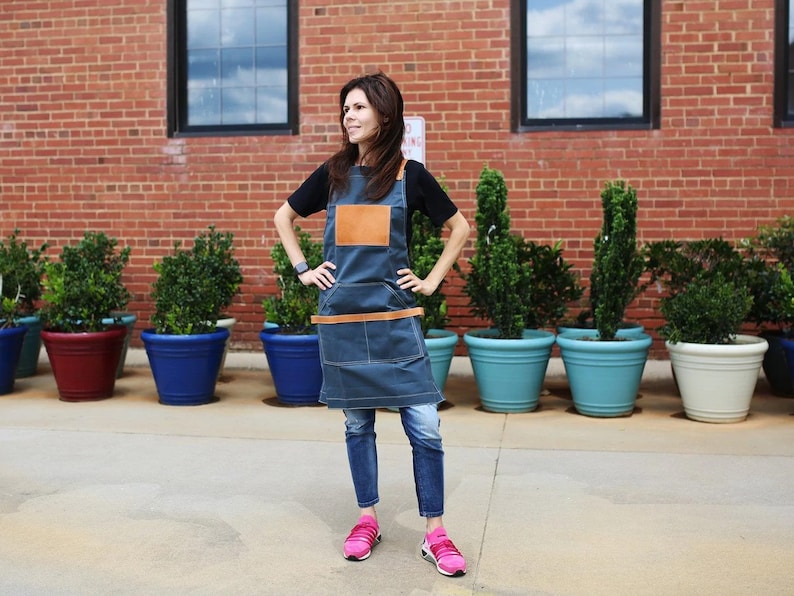 May include: A woman wearing a blue denim apron with a brown leather patch and a brown leather belt. She is standing in front of a brick wall with plants in pots.